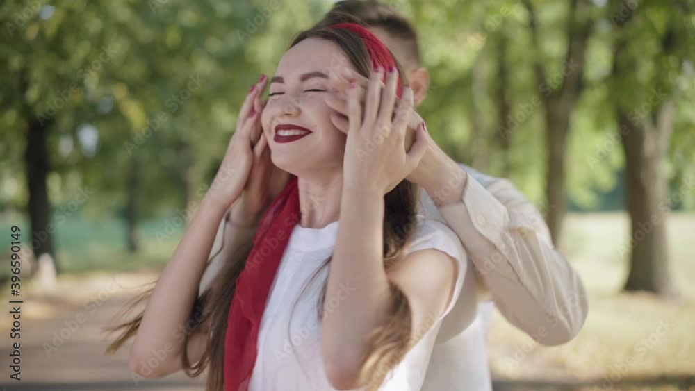 Cheerful young man closing eyes of beautiful young woman waiting for a date and hugging adorable partner. portrait of happy Caucasian boyfriend and girlfriend meeting in sunny summer park.