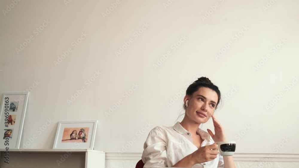 A smiling young woman wearing earbuds is drinking a cup of coffee while talking to someone sitting in the room A smiling young woman wearing earbuds is drinking a cup of coffee while talking to someone sitting in the room