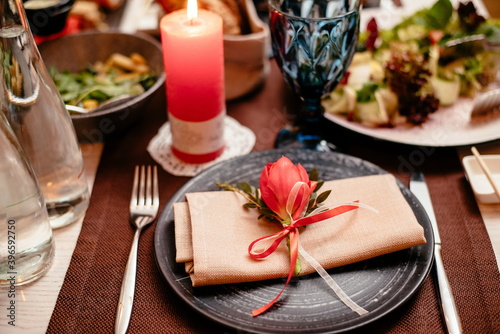 Empty black ceramic plate with a knife, fork on a napkin on a brown background, decorated with a red tulip, close-up. Elegant stylish table setting.