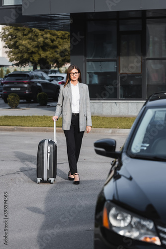  businesswoman walking with suitcase near black car on blurred foreground