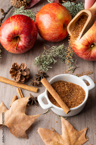 Top view of small white bowl with brown sugar  with falling sugar  red royal gala apples with cinnamon sticks and autumn leaves  with selective focus  on wooden table  vertical