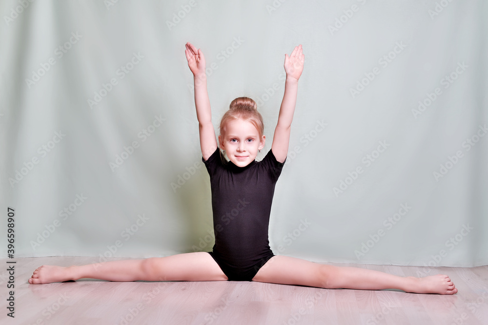 Gymnast girl sitting do the splits inside the room, in a black swimsuit.