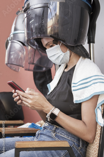 one hispanic young adult woman sitting in a hair dryer hood beauty salon or spa using protective mask and using smartphone during coronavirus covid-19 pandemic