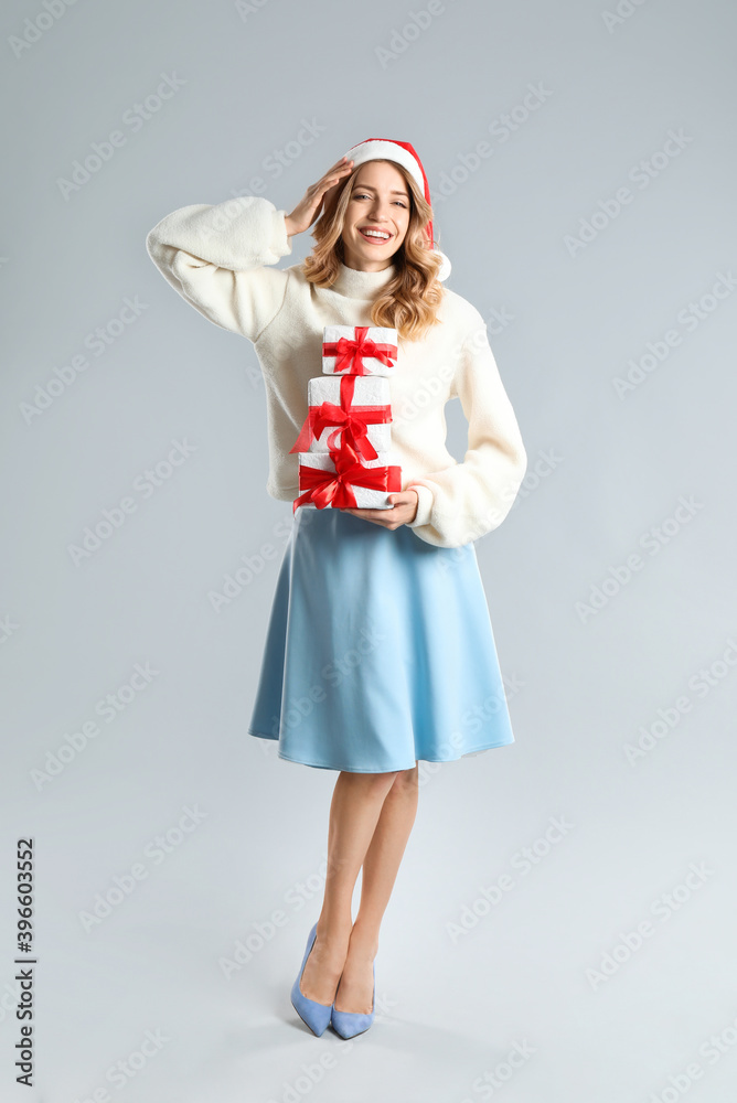 Beautiful young woman in Santa hat with Christmas presents on light grey background Beautiful young woman in Santa hat with Christmas presents on light grey background