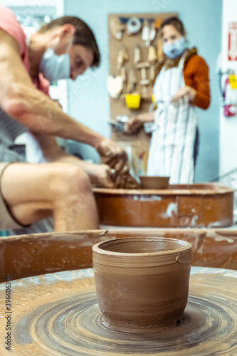 Bowl of raw clay dries on a potter's wheel against the background of a man working in a pottery workshop.