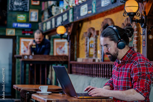Young bearded freelancer or blogger, sitting in a pub, working on a laptop computer, wearing earphones on his head. Another customer having a drink at an appropriate distance. Social distancing. 