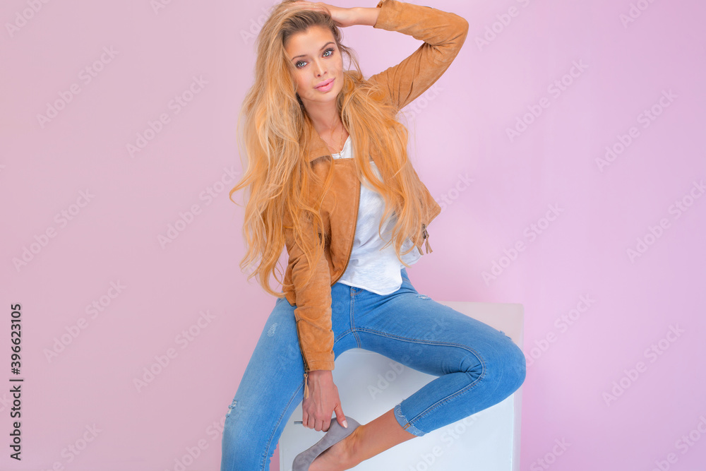 Beautiful sexy blonde girl with long hair and tanned body wearing brown jacket and blue jeans, sitting and sensual posing at pink wall.