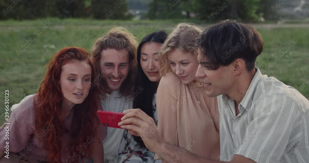 Crop view of young people using smartphone frontal camera while sitting on grass. Diverse happy friends smiling while taking selfie and looking at photo. Concept of friendship Crop view of young people using smartphone frontal camera while sitting on grass. Diverse happy friends smiling while taking selfie and looking at photo. Concept of friendship