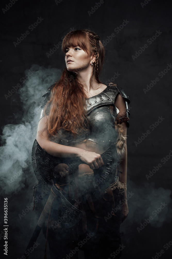 Portrait of warlike female fighter from north with long brown hairs in dark armour with two handed axe in dark smokey background.