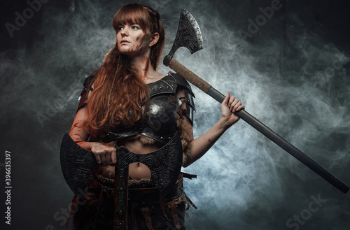 Violent and warlike northern woman warrior holding axes and posing in dark smokey background. © Fxquadro