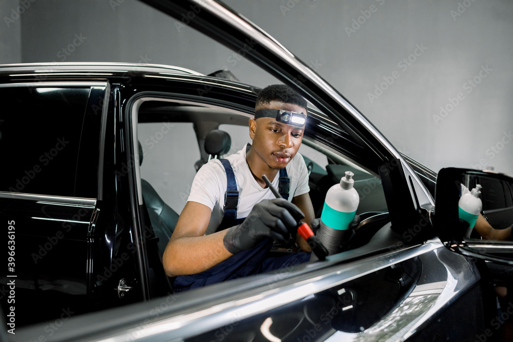 Front view of handsome African man worker of car detailing workshop, wiping car door leather panel with soft brush and special professional anti scratch and care cream milk