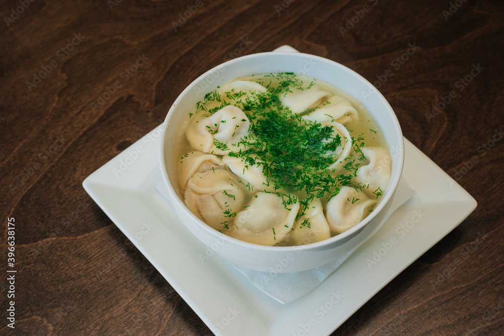 Dumplings with meat in broth with herbs in a white plate on a dark background. Russian folk cuisine. White bowl on a wooden table. Dumplings with meat in broth with herbs in a white plate on a dark background. Russian folk cuisine. White bowl on a wooden table.