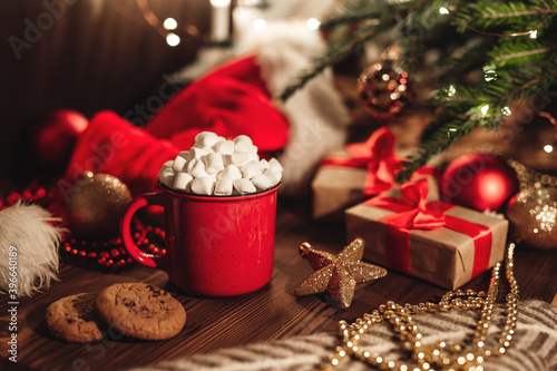 Christmas red mug with cocoa and marshmallows and cookies on a wooden table. New Year's still life with a Christmas tree, a Santa hat and festive decorations.