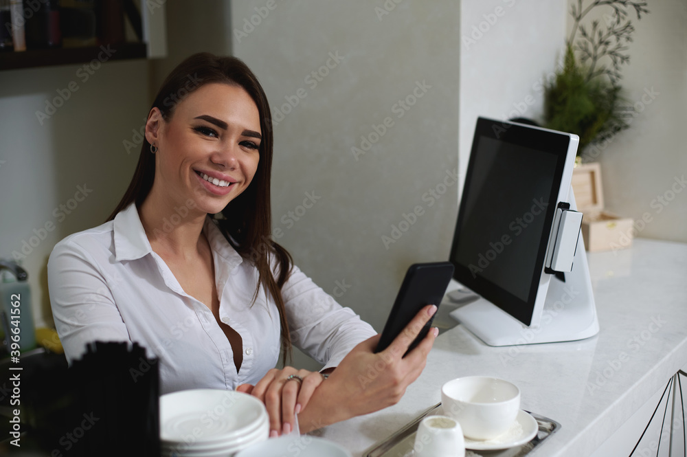 Cute brunette behind the bar smiles at the camera and holds the phone in her hands Cute brunette behind the bar smiles at the camera and holds the phone in her hands