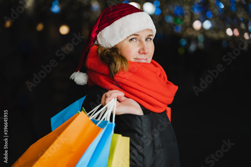 Christmas shopping of young beautiful woman. Colorful bright paper bags with gifts,presents.Walking on market street in city.Garlands,lights.Stylish look, jacket, santa claus hat.New year celebration