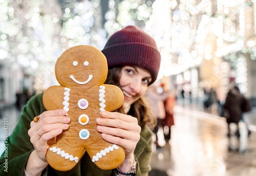 Young beautiful girl, woman with huge funny gingerbread man. Christmas shopping. Walking on market street in big city decorated with garlands, lights.Stylish look, wool coat, hat.New year celebration photo