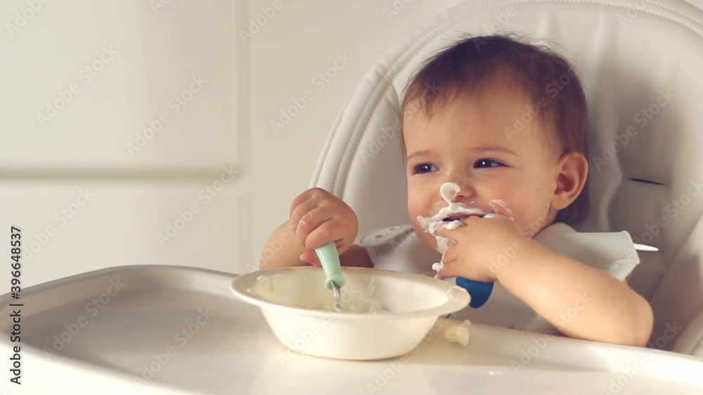 Cute little baby girl eating baby food with spoon. her face is smeared with yoghurt on high chair in home kitchen Cute little baby girl eating baby food with spoon. her face is smeared with yoghurt on high chair in home kitchen