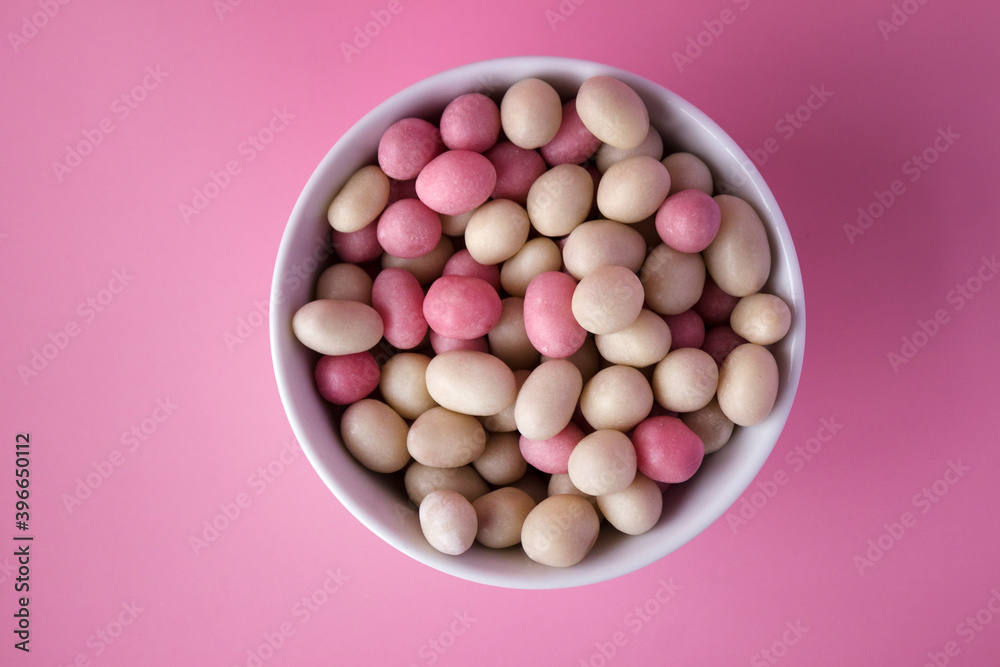White and pink round candies in glaze and with peanuts inside are laid in plate on a pink background. View from above. White and pink round candies in glaze and with peanuts inside are laid in plate on a pink background. View from above.