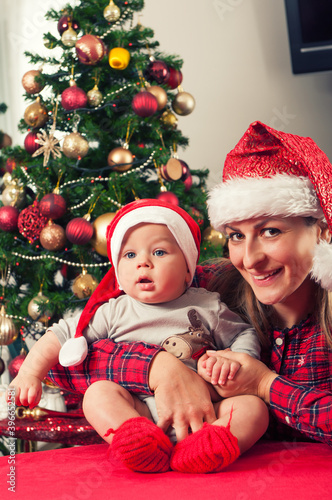 Mother and her beautiful baby boy posing beside christmass tree with santa claus hats