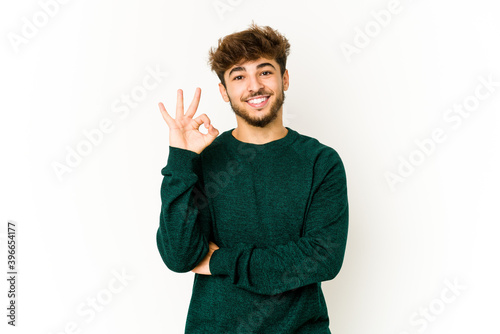 Young arab man on white background winks an eye and holds an okay gesture with hand.