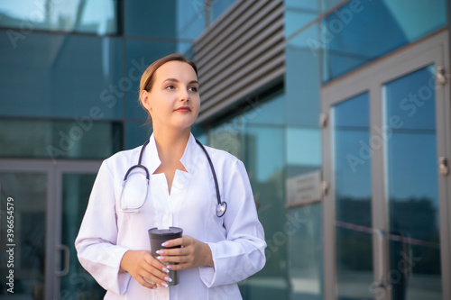 A few minutes to relieve stress in fresh air to start treatment for following patients. Young female doctor holds cup of tea during her break and looking away. Medicine and healthcare concept.