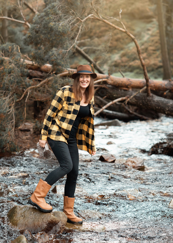 Happy woman in yellow shirt and hat walking along the river in the autumn forest © Iryna