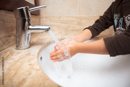 Young kid rinsing off soap from his hands under tap water after washing them during pandemia as learned in school and at home with rolled-up sleeves in a sink surrounded by brownish marble