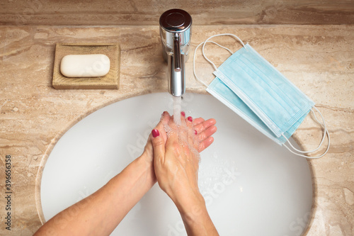 Young woman washing her hands and rinsing off the soap under the tap water, two new surgical masks ready on her right during pandemic