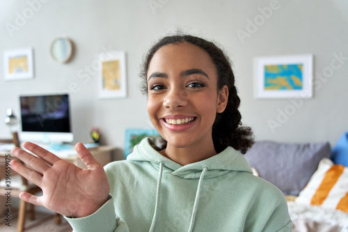 Happy african gen z teen girl waving hand talking to camera in bedroom. Mixed race teenager recording vlog, streaming for social media, video calling in online chat at home. Web cam view headshot photo