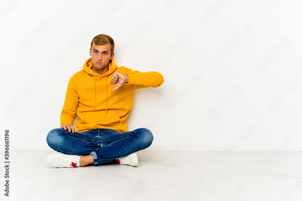 Young caucasian man sitting on the floor showing thumb down, disappointment concept. Young caucasian man sitting on the floor showing thumb down, disappointment concept.