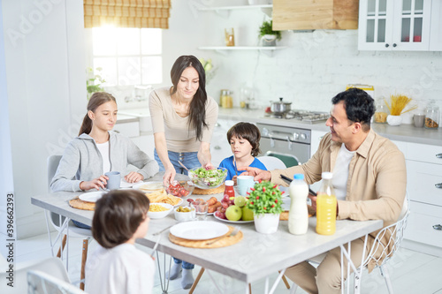 Share food. Caring hispanic woman serving salad for her husband and children, standing in the kitchen. Latin family having dinner together at home