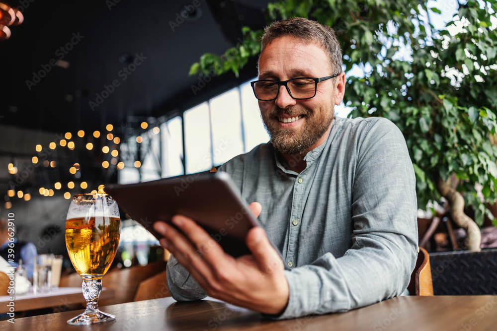 Smiling middle aged bearded man with eyeglasses sitting in a bar after work and using tablet for hanging on social media. On a table is a fresh cold light beer. Smiling middle aged bearded man with eyeglasses sitting in a bar after work and using tablet for hanging on social media. On a table is a fresh cold light beer.