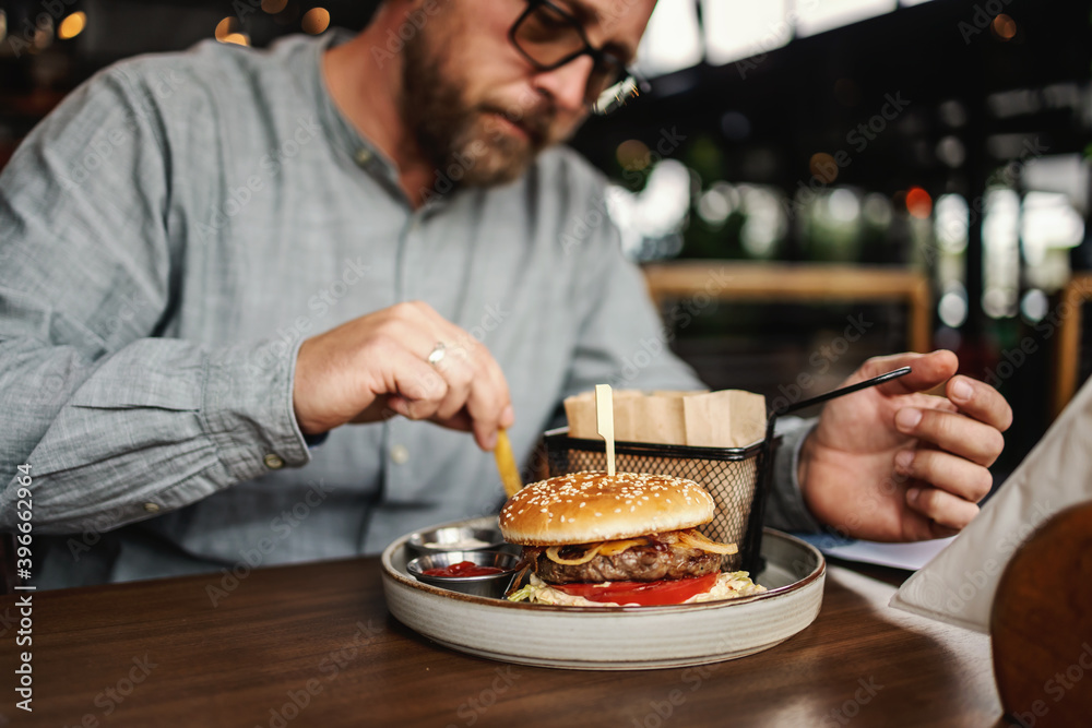Middle aged bearded hungry man sitting in restaurant and eating delicious burger.