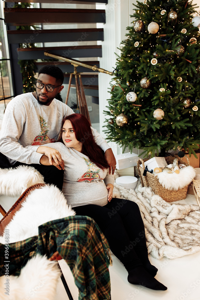 Loving African American husband gently hug lovely pregnant wife, wonderful couple sit near Christmas tree at home, enjoy tenderness, cozy winter holidays, xmas celebration concept