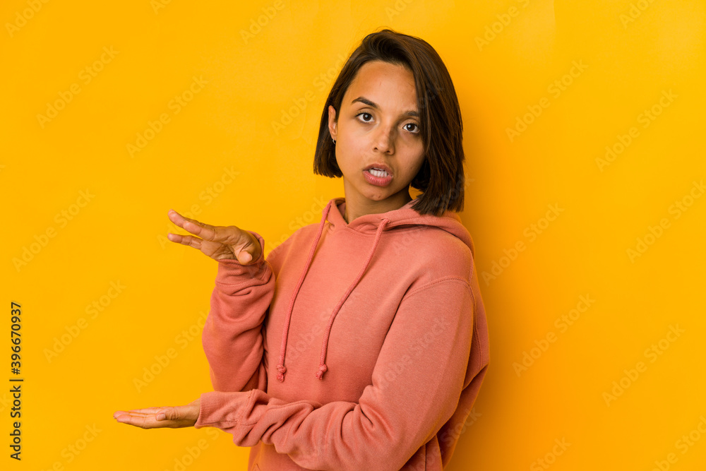 Young hispanic woman isolated on yellow shocked and amazed holding a copy space between hands. Young hispanic woman isolated on yellow shocked and amazed holding a copy space between hands.