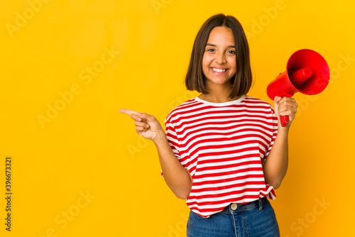 Young hispanic woman holding a megaphone smiling and pointing aside  showing something at blank space.