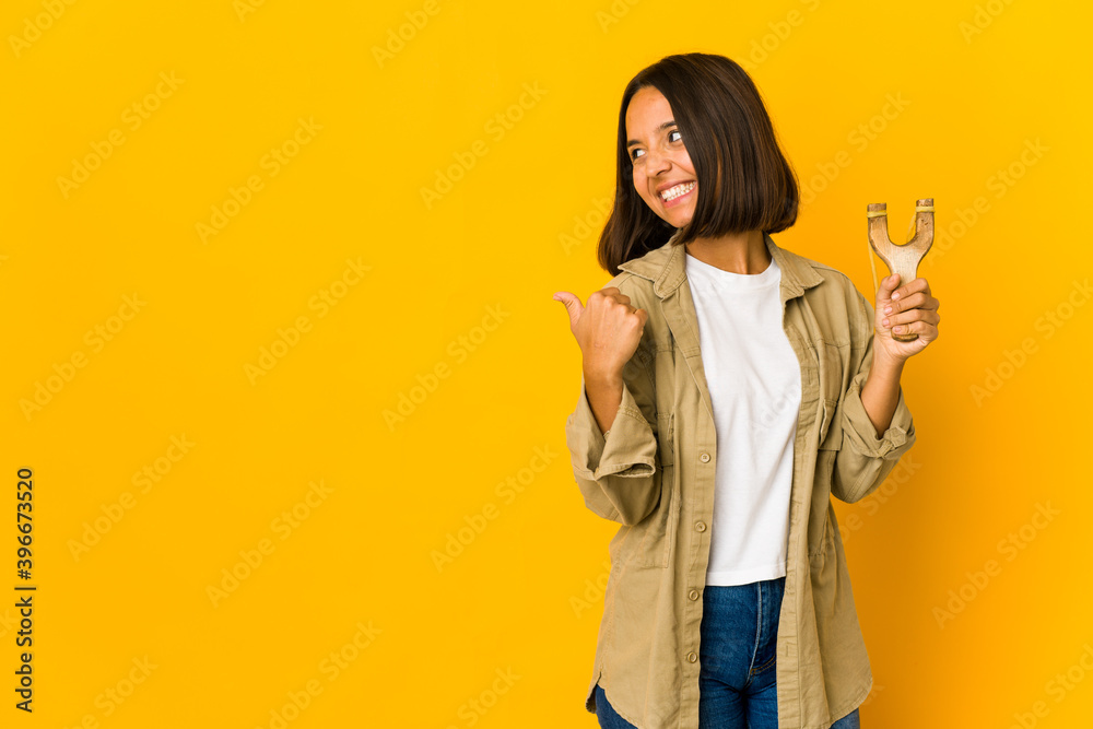 Young hispanic woman holding a slingshot points with thumb finger away, laughing and carefree. Young hispanic woman holding a slingshot points with thumb finger away, laughing and carefree.