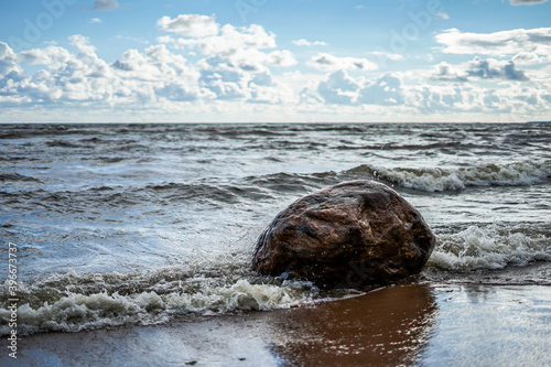 The wave beats against a large stone on the seashore, a clear day, light clouds