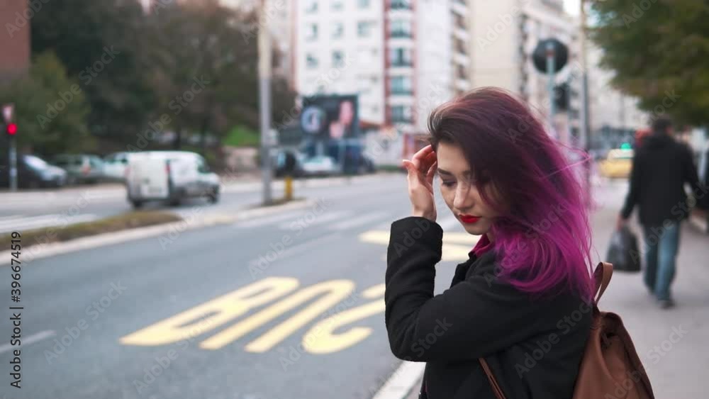 Pretty asian hipster girl waiting at a bus stop on a busy street. Woman tourist at sidewalk. Concept of lifestyle, travel and tourism.Pink colored hair charming millenial using public city transport. Pretty asian hipster girl waiting at a bus stop on a busy street. Woman tourist at sidewalk. Concept of lifestyle, travel and tourism.Pink colored hair charming millenial using public city transport.