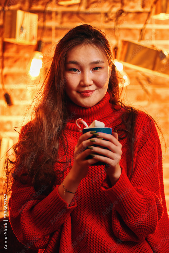 Asian girl in sweater with cup of hot cocoa. Coffee with marshmallows and lollipops. Christmas atmosphere. New year mood. Romantic portrait. Light bulbs and present boxes background of brick wall.
