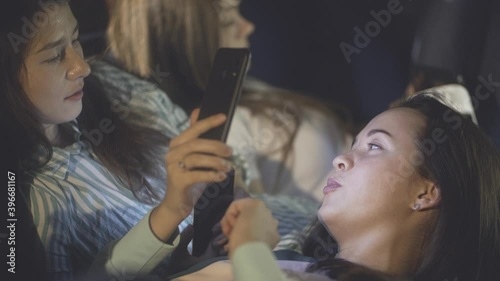 Homosexual multiracial couple lying on sofa at home party in evening or night. Happy millennials fubbing during friendly meeting. Girls connecting and discussing news on Internet. Spend time together. photo