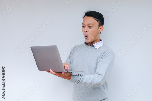 Wow face of Young Asian man shocked what he see in the laptop when working isolated grey background wearing grey shirt