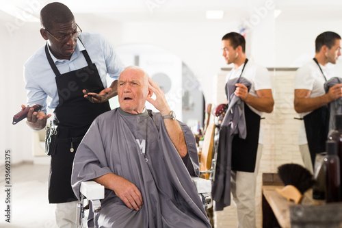 Portrait of upset dazed aged man sitting in barber chair with confused African hairdresser behind him