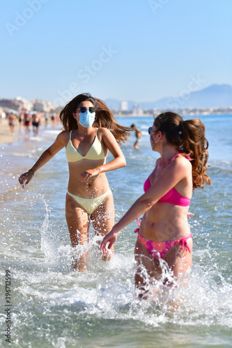 Women wearing face masks, glasses and bikinis running on water at the sea on an out of focus background. Safety and new holidays concept.