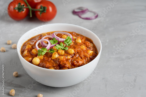 Chole masala or chana indian vegan food made of cooked chickpeas, tomatoes and cumin decorated with onion rings and parsley served in white bowl on gray concrete background. Image with copy space