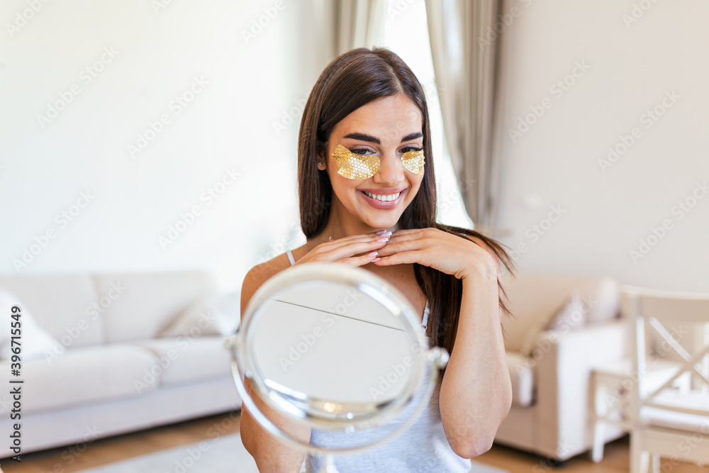 Portrait of pretty, nice young woman with golden eye patches, having bags under eyes in the morning because of less sleep, showing an effect of perfect skin. Looking in the mirror Portrait of pretty, nice young woman with golden eye patches, having bags under eyes in the morning because of less sleep, showing an effect of perfect skin. Looking in the mirror
