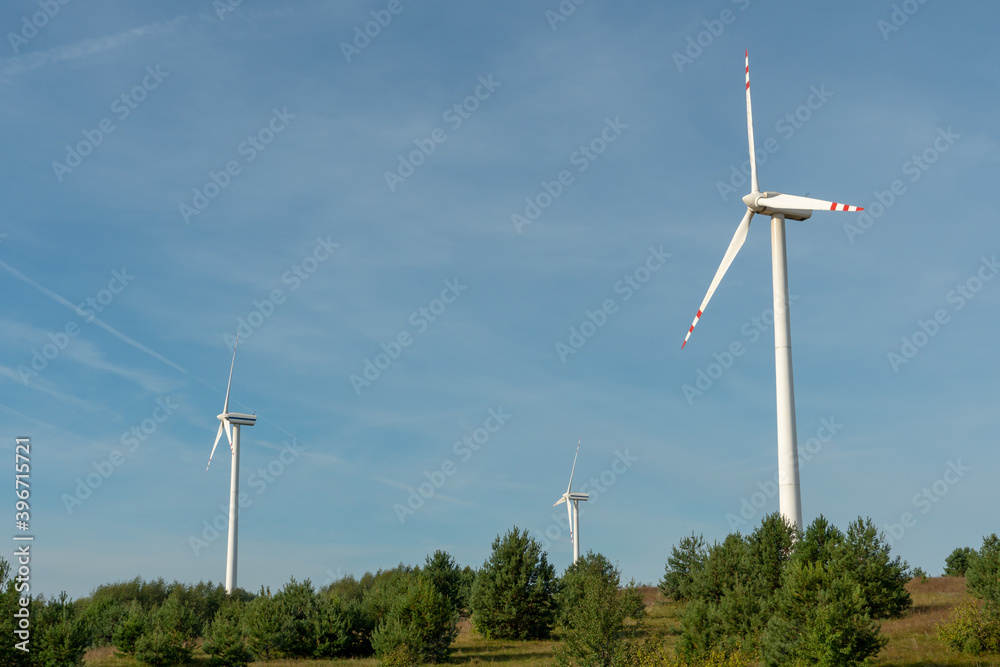 view of a modern windmill against a blue sky. The white blades of the wind turbine. Wind turbines in the forest away from the big city on a summer day. Clean and renewable energy production