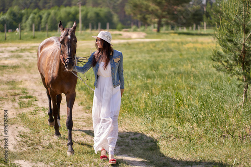 A woman in a straw hat and white dress leads a horse in the pasture.