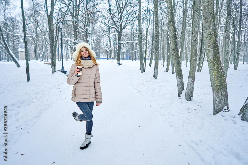 Snowy winter in the park. A woman in a jacket and a fur hat is having fun in front of the camera. Paper cup for coffee in hands.