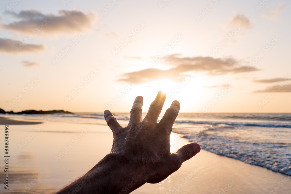 Male hand trying to touch the sun at the beach shore by sunset - Reaching, peace and hope concept - Jarugo beach - Fuerteventura